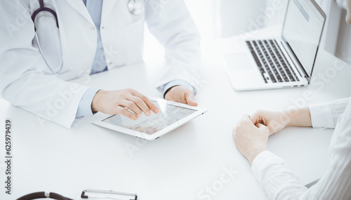 Doctor and patient sitting opposite each other at the desk in clinic. The focus is on female physician's hands pointing into tablet computer touchpad, close up. Medicine concept