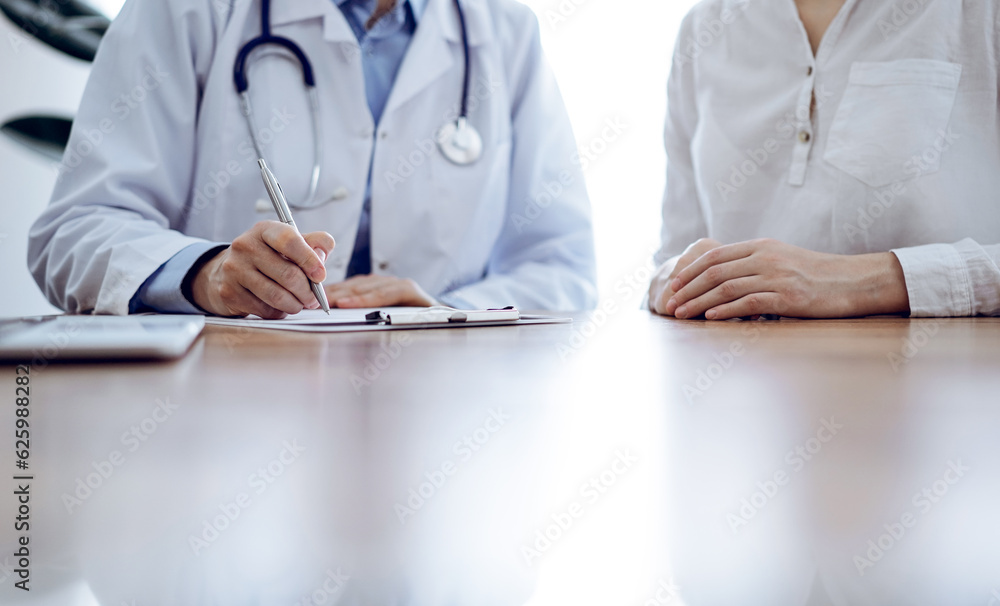 Doctor and patient sitting near each other at the wooden desk in clinic. Female physician's filling up a records form. Medicine concept Doctor and patient sitting near each other at the wooden desk in clinic. Female physician's filling up a records form. Medicine concept
