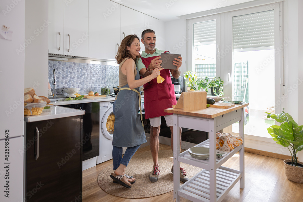 wide shot of an adult caucasian couple in the middle of the kitchen. They are standing in their aprons smiling and looking at a recipe on their tablet wide shot of an adult caucasian couple in the middle of the kitchen. They are standing in their aprons smiling and looking at a recipe on their tablet