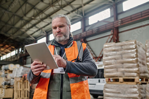 Mature foreman in reflective clothing using tablet pc to control the number of cargo in warehouse