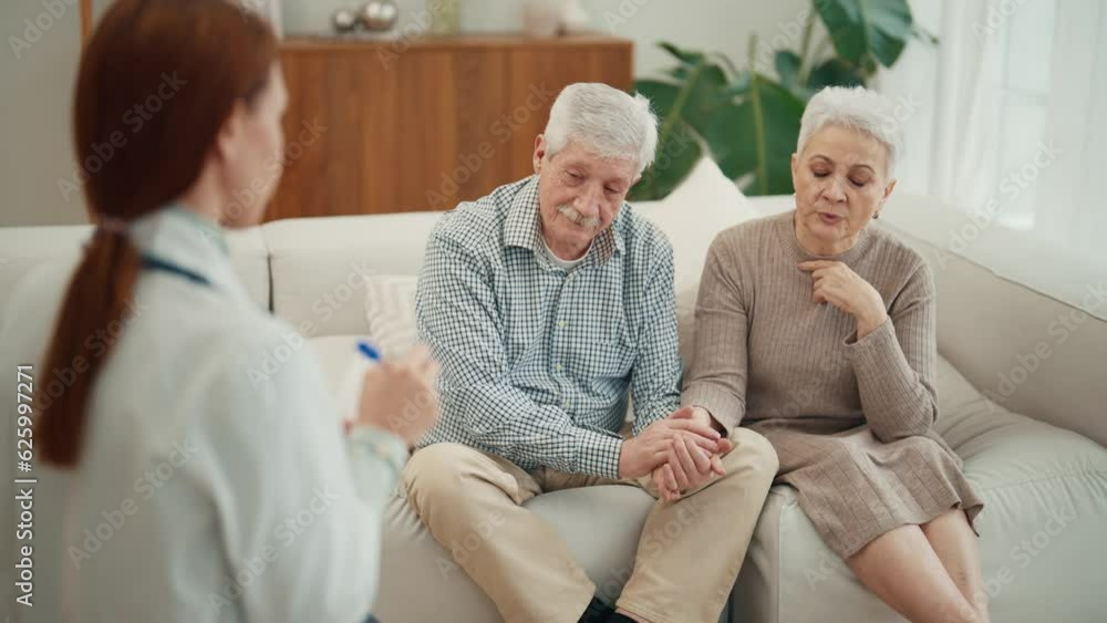 Young woman doctor nurse social worker visits senior couple. Old woman complaints about her health sits on couch with husband at home. Family support, relationship in old age and medical help concept. Young woman doctor nurse social worker visits senior couple. Old woman complaints about her health sits on couch with husband at home. Family support, relationship in old age and medical help concept.
