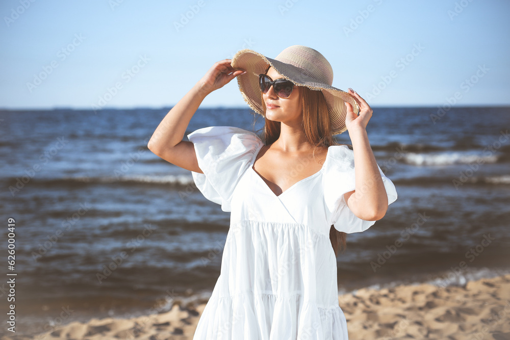 Happy blonde woman is posing on the ocean beach with sunglasses and a hat. Evening sun