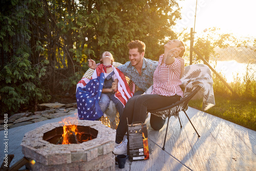 Joyful man and woman with daughter sitting by fireplace outdoors, roasting marshmallows and laughing, having a good time together.