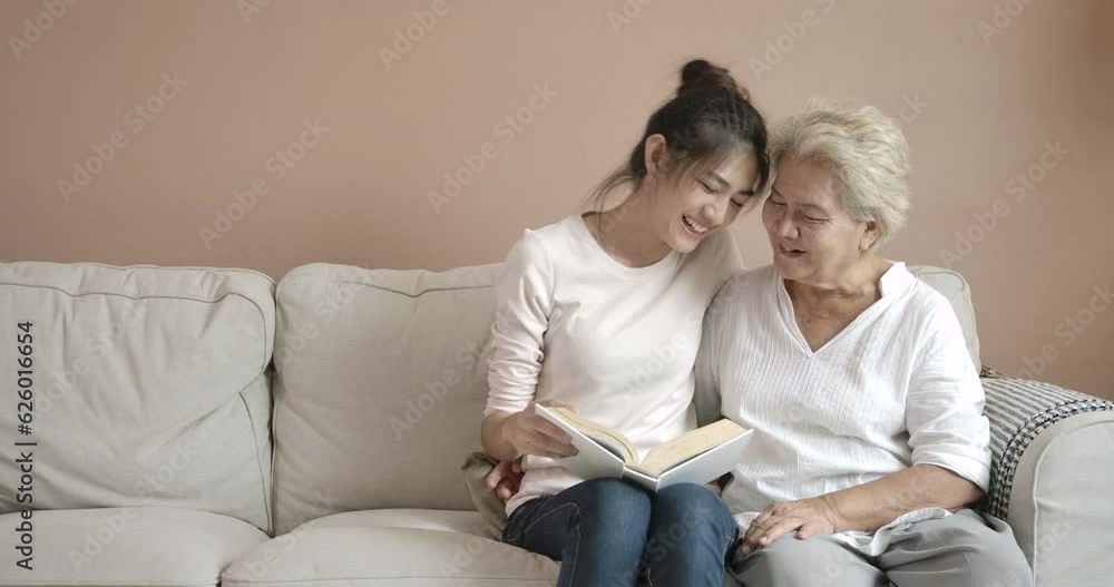 Young beautiful asian woman and her mother reading book together on sofa. Happy mom and pretty daughter in casual clothes at house during quarantine. Social distance new normal concept. Young beautiful asian woman and her mother reading book together on sofa. Happy mom and pretty daughter in casual clothes at house during quarantine. Social distance new normal concept.