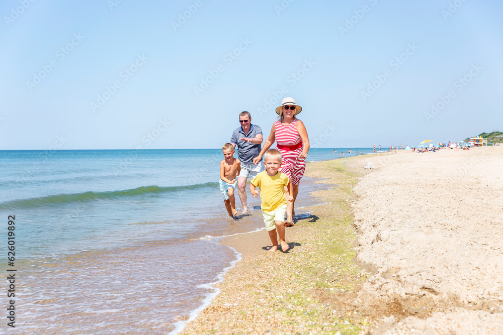 Elderly man and woman play with children on the seashore. Joyful grandparents run after their grandchildren along the sandy beach on a sunny day. Love and tenderness. Travel and active lifestyle.
