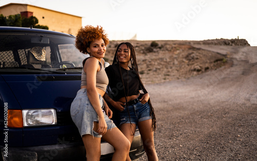 Two cute girls with dark skin are posing in the front of a camper van. The woman with the braided hair looks happily at the woman with the afro hair who looks at the camera. Trips of African women.