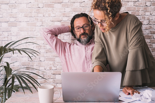 Two colleagues man and woman working together with computer at office. Small business people lifestyle concept. Entrepreneur and explanation. Mature freelance male stretching arms and female working photo