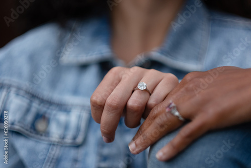 Showing off their engagement rings, a lesbian celebrates their love and their upcoming wedding. They are both wearing jeans, and are Black and multiracial.