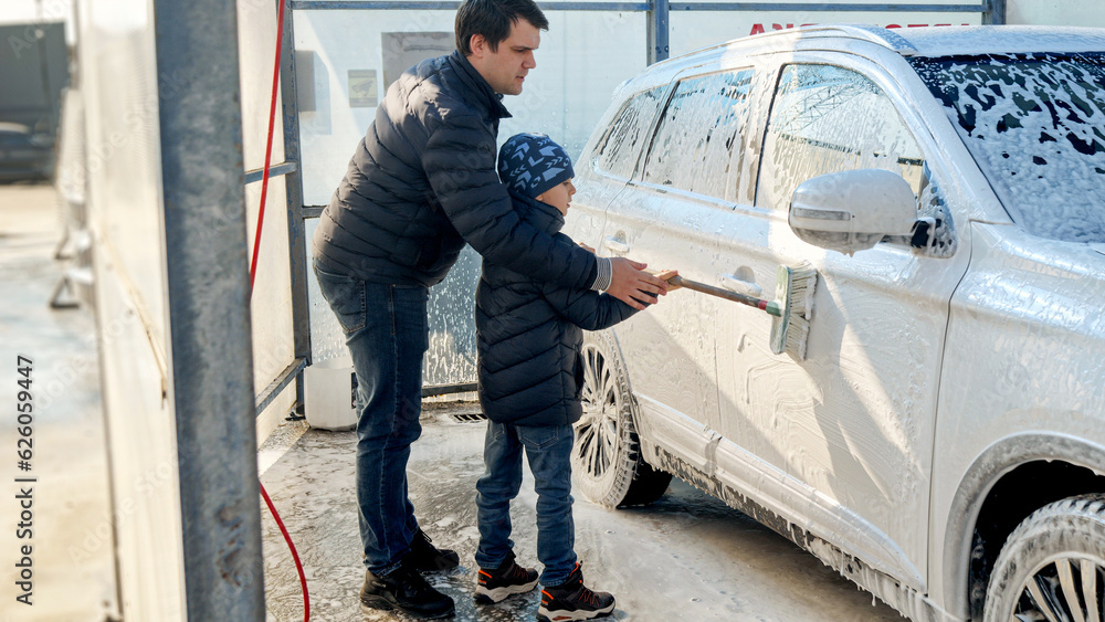 Young smiling man with son washing car outdoors with brush and foam. Automobile care, transport cleaning, dirty car.