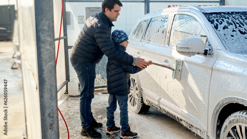 Young smiling man with son washing car outdoors with brush and foam. Automobile care, transport cleaning, dirty car. Young smiling man with son washing car outdoors with brush and foam. Automobile care, transport cleaning, dirty car.