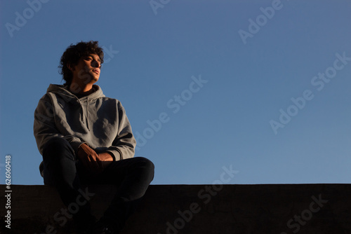 reflective young man sitting alone on the rooftop of a building gazes at the horizon and contemplates the sunset.