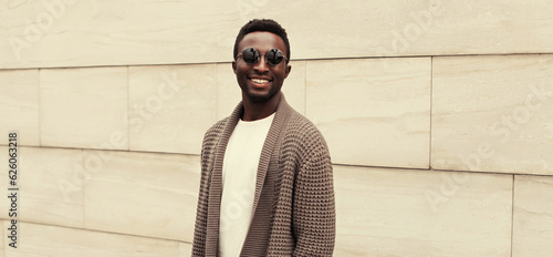 Portrait of stylish smiling young african man wearing brown knitted cardigan on city street background