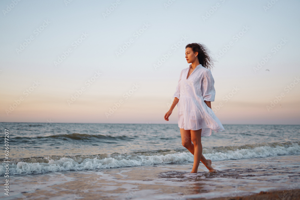 Slender young woman in airy white is enjoying the sunset light on the seashore. Summer time. Travel, weekend, sunny, fun, positive mood. Active lifestyle.