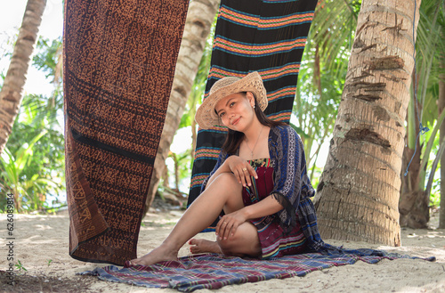 A young Asian woman is sitting while wearing clothes made of woven cloth combined with batik cloth with a tree in the background