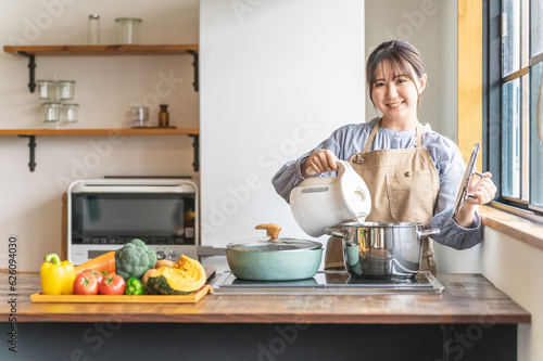 キッチンで電気ケトルのお湯を鍋に入れるエプロン姿のアジア人女性
