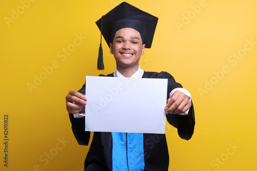 Obraz na plátne Excited graduate young man in mortar board and bachelor gown holding blank poster with copy space on yellow studio background