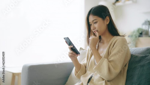 Happy young asian woman relax on comfortable couch at home, typing chat message on smartphone, smiling girl use cell phone chatting, searching information on browser wireless internet, online shopping