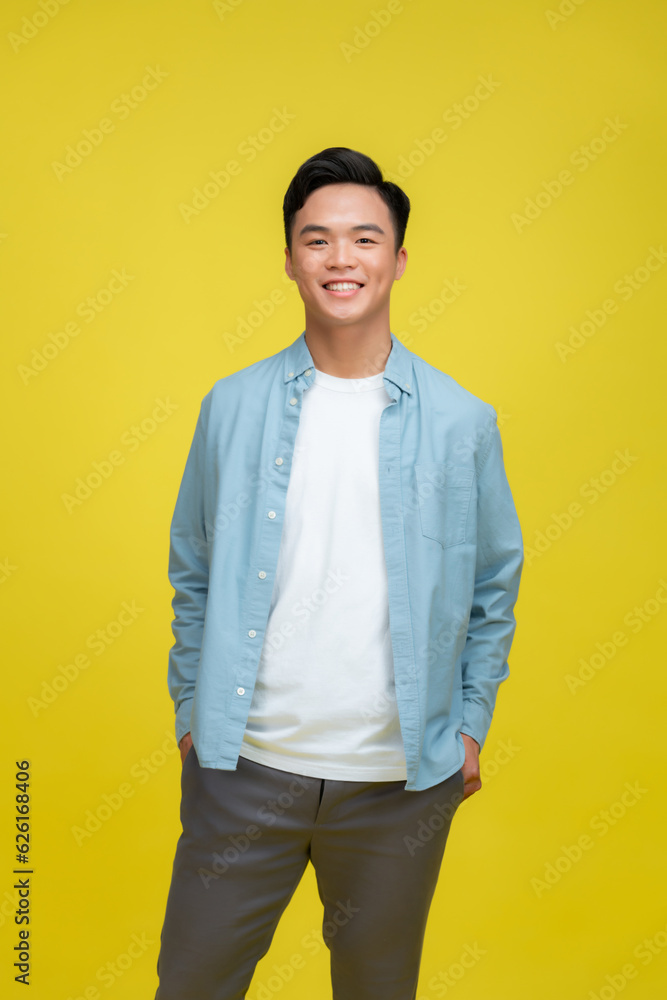 smiling young handsome Asian man standing with one hand in pocket isolated on yellow studio background smiling young handsome Asian man standing with one hand in pocket isolated on yellow studio background