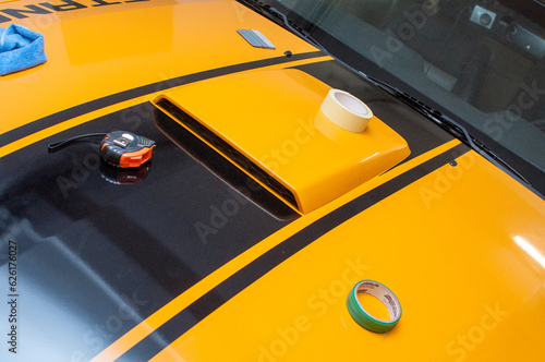 Installation (gluing, applying) of black strips decal of glossy foil on a yellow classic sports car in a garage.