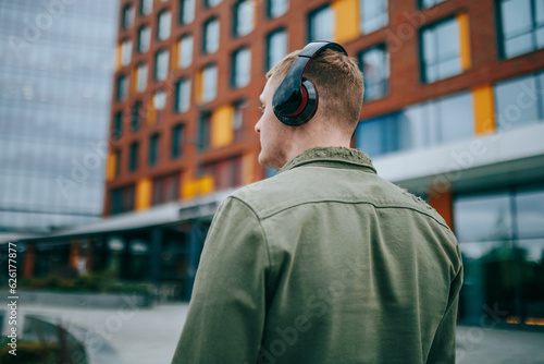 A caucasian man with headphones around his head, seen from the back, enjoying the beautiful architecture of the city while listening to music. © Alexandr