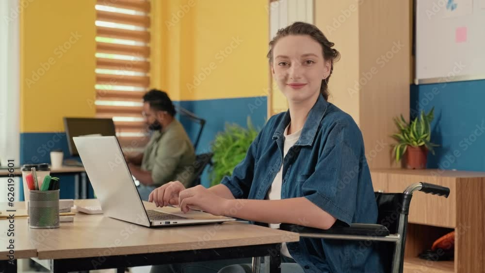 Portrait of young woman in wheelchair working on laptop and looking at camera at office Portrait of young woman in wheelchair working on laptop and looking at camera at office