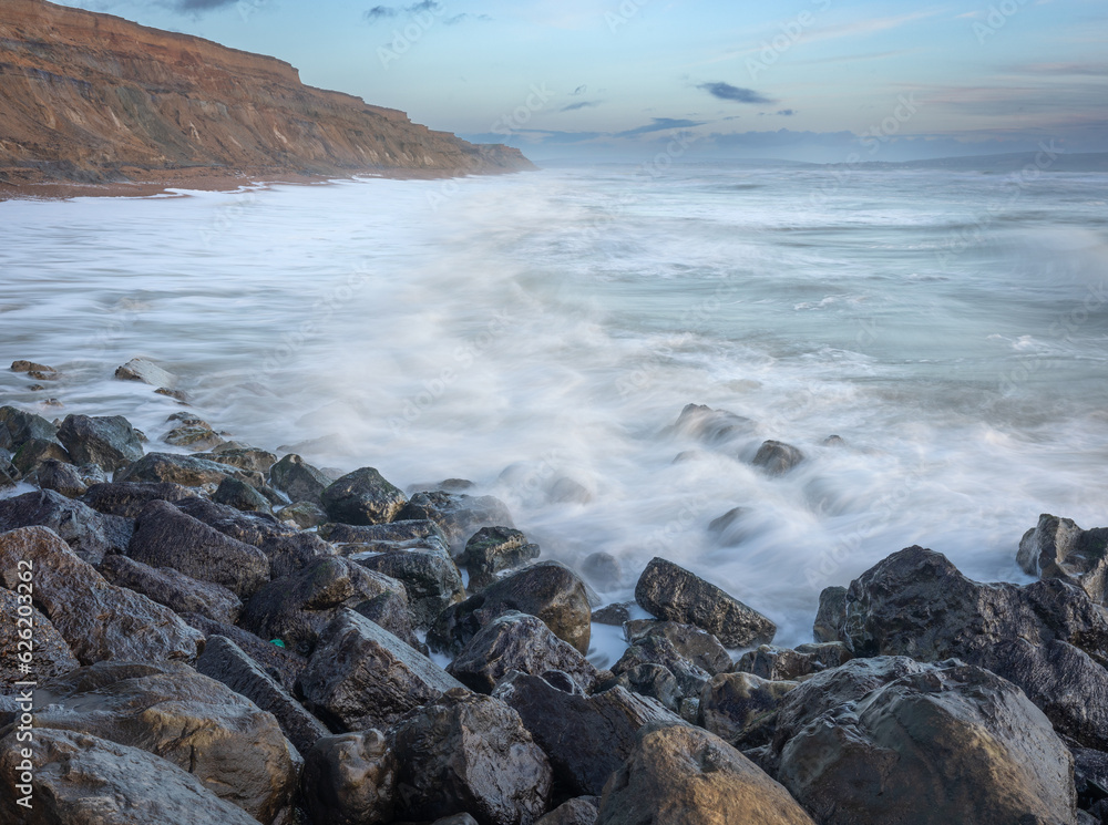 An early morning seascape scene with shiny rocks in the foreground, blurred waves over rocks and high sand stone cliffs going into the distance