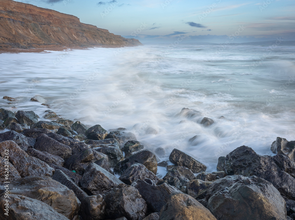 An early morning seascape scene with shiny rocks in the foreground, blurred waves over rocks and high sand stone cliffs going into the distance An early morning seascape scene with shiny rocks in the foreground, blurred waves over rocks and high sand stone cliffs going into the distance