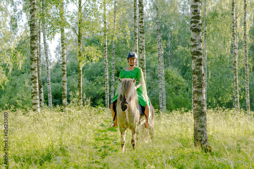 Barefoot woman rider on Icelandic horse in sunny Finnish brirch  forest during sunset. Fairytale like feeling. Rider is wearing helmet.
