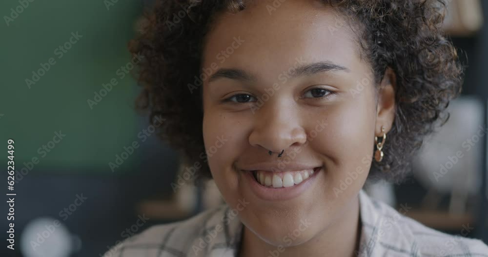 Close-up slow motion portrait of happy African American girl smiling looking at camera against blurred indoor background. People and emotion concept. Close-up slow motion portrait of happy African American girl smiling looking at camera against blurred indoor background. People and emotion concept.