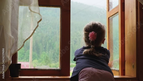 Child opening window while raining outdoors at the mountains. Child sitting in the house and looking outdoors, waiting for nice weather, observing nature, thinking. Relaxed sad kid alone at home