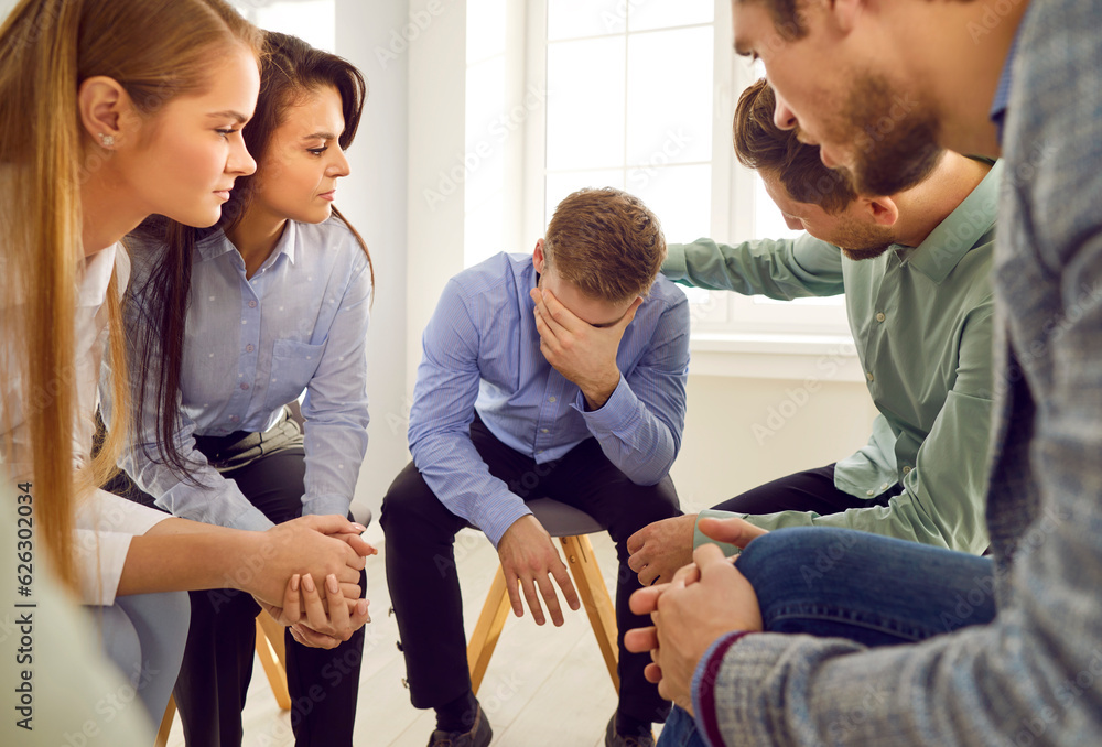 Obraz premium Team of colleagues, friends, or people in group therapy sitting in a circle and trying to support and comfort a sad young man who is hiding his face and crying. Support concept