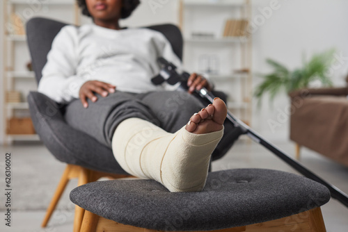 African american woman is sitting at home in armchair with broken bandaged leg lying on stool and holding crutches in her hands. Recovery time after physical injury of the foot bone. Selective focus.