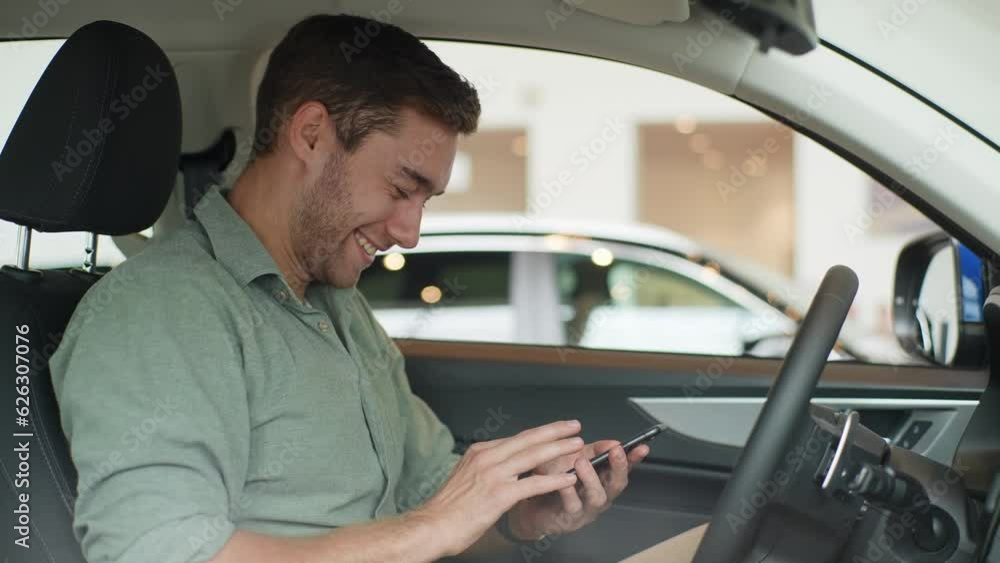 Side view portrait of cheerful bearded male driver sitting behind wheel of car, using smartphone, laughing looking at the screen. Happy man texting on internet on cell phone laughing at message. Side view portrait of cheerful bearded male driver sitting behind wheel of car, using smartphone, laughing looking at the screen. Happy man texting on internet on cell phone laughing at message.