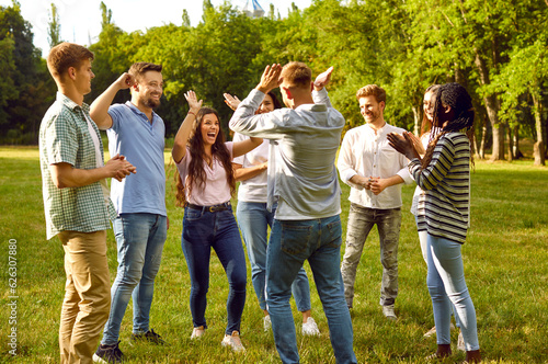 Large group of fun cool young best friends hanging out together in park in summer. Male and female friends laughing giving each other high five in nature against background of green trees.