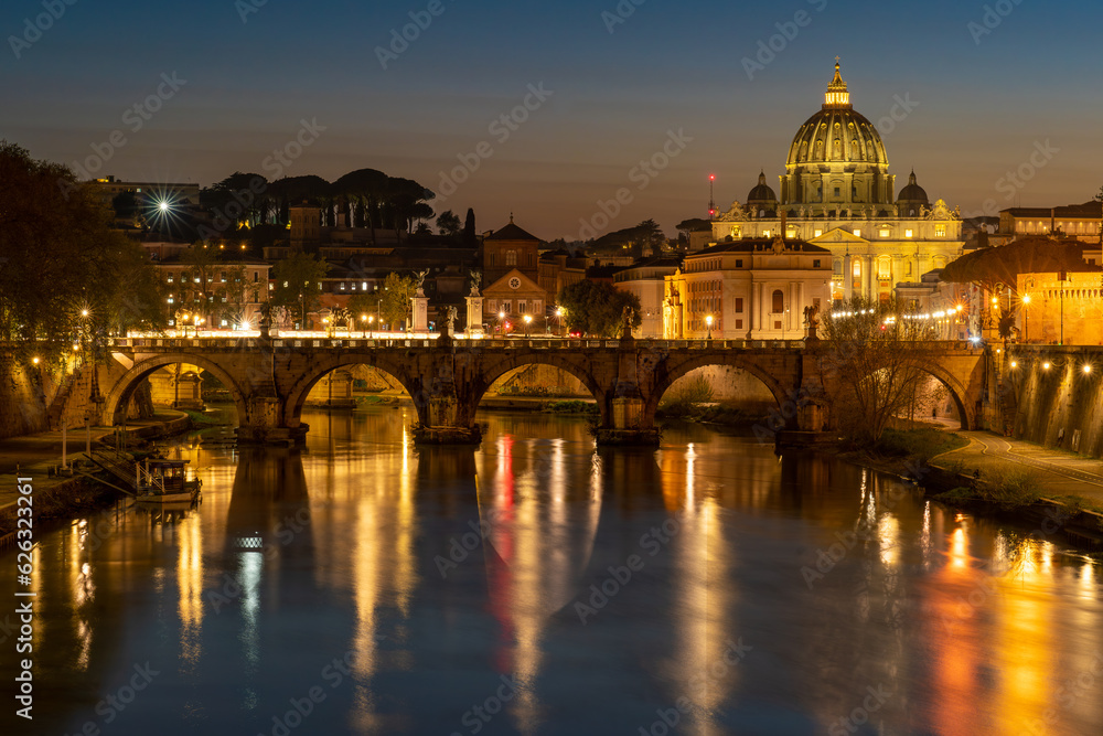 Nighttime photo of the Ponte Umberto 1 Bridge over the Tiber River at Night in Rome, Italy. St. Peter's Basilica is visible in the background and bathed in golden light.