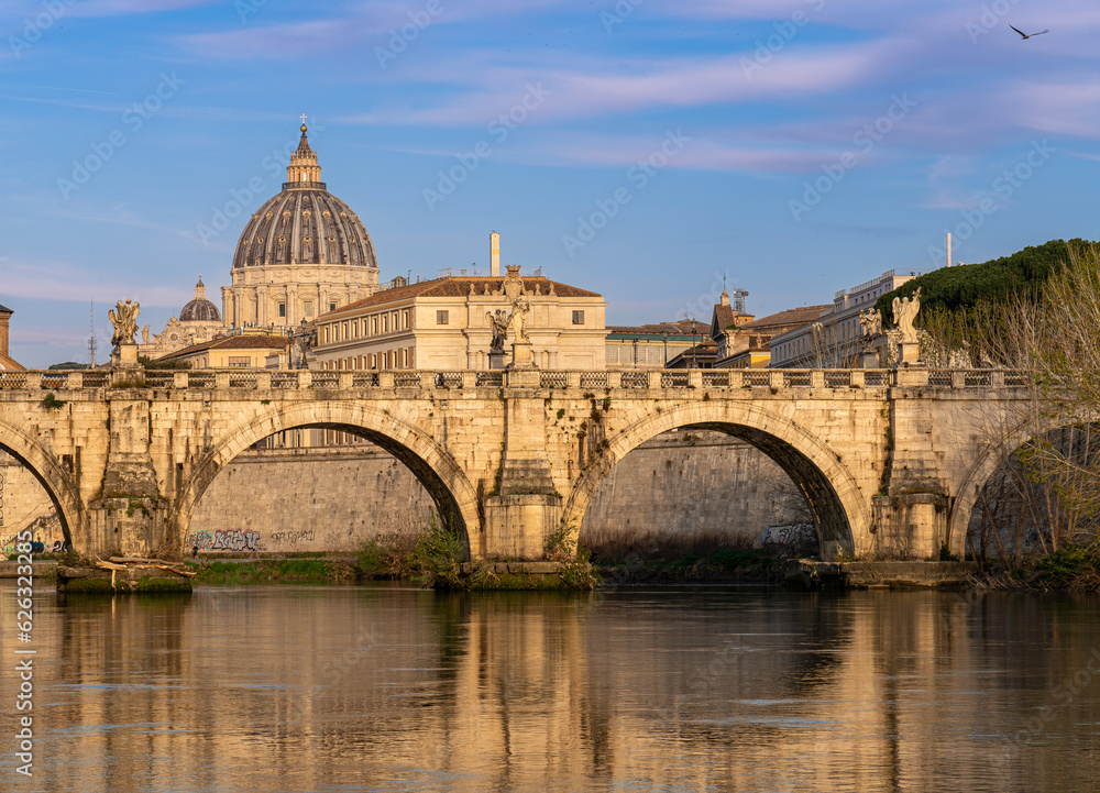 Daytime photo of the Ponte Umberto 1 Bridge over the Tiber River at Night in Rome, Italy. St. Peter's Basilica is visible in the background and both are bathed in golden sunrise light.