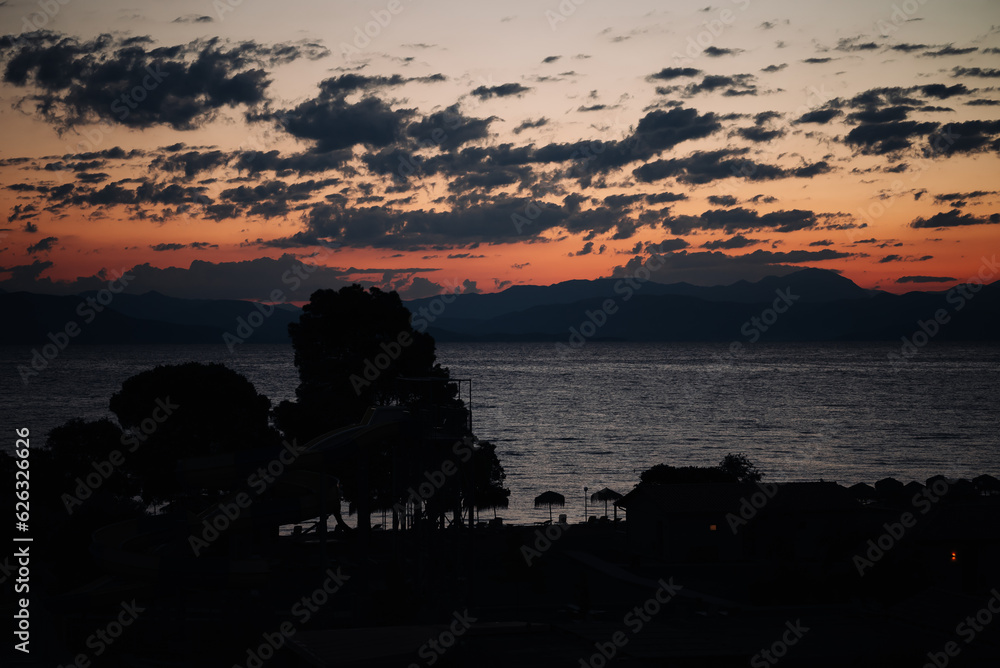 Sunrise From Corfu Island Overlooking Mountains Of Balkan Peninsula, Corfu. In Foreground Are Silhouettes Of Trees And Water Slide. Concept Of Influence Of Sun On Human Body.