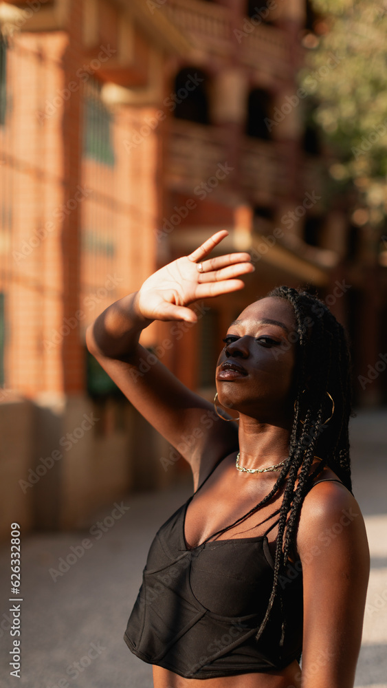 Obraz premium A young black woman with braids in her hair shields her eyes from the sun with her hand during a walk in the city