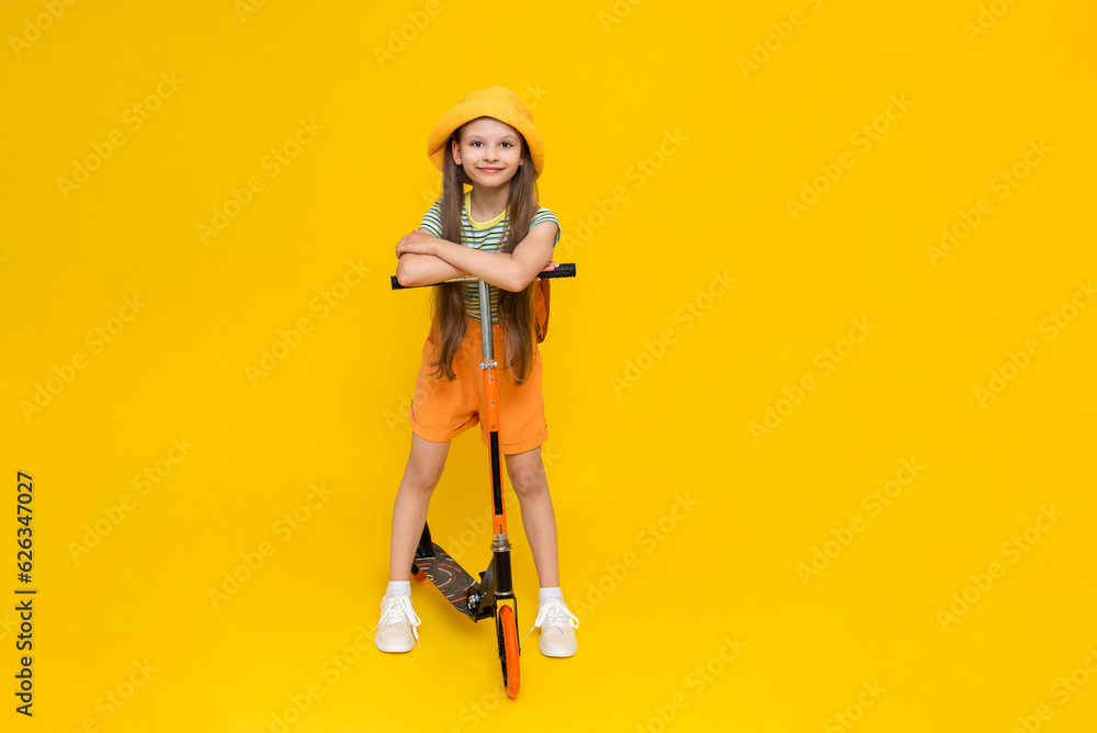 A full-length happy, cheerful young girl in a hat and shorts riding a two-wheeled scooter. Children's active city recreation. Bright yellow isolated background. A full-length happy, cheerful young girl in a hat and shorts riding a two-wheeled scooter. Children's active city recreation. Bright yellow isolated background.