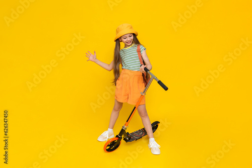 A full-length young girl in a hat and shorts is standing next to a scooter and enjoying a summer vacation. Active city recreation for children. Yellow isolated background.