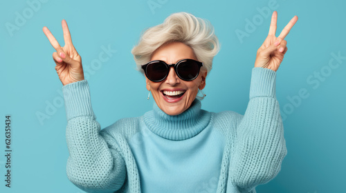 Elderly stylishly dressed woman showing peace sign on fingers on blue background.