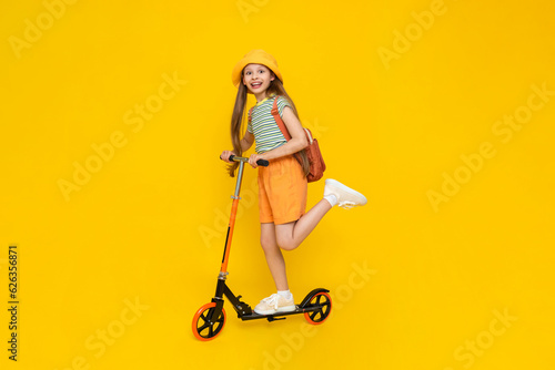 A child rides a scooter. A full-length side view of a beautiful cheerful dreamy young girl. Bright yellow isolated background.