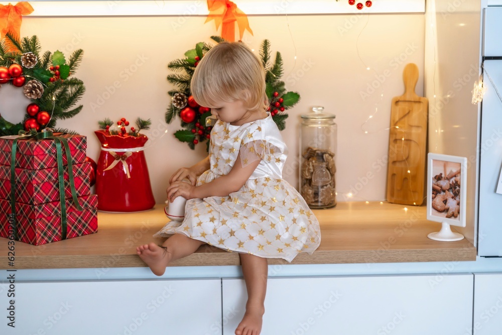 Christmas kitchen girl. Portrait of a charming little girl 2 years old, she sits on a table in a white dress and drinks from a red mug in a decorated kitchen for christmas. Christmas kitchen girl. Portrait of a charming little girl 2 years old, she sits on a table in a white dress and drinks from a red mug in a decorated kitchen for christmas.
