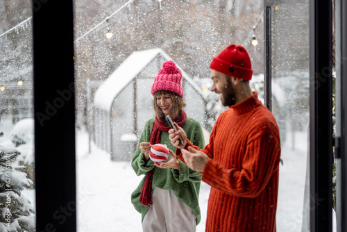 Man and woman prepare to decorate Christmas tree with festive balls, standing together happily at snowy backyard. Young family celebrating winter holidays at home