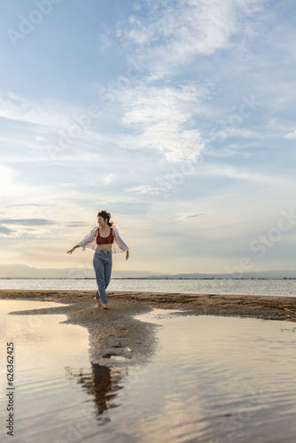 woman on the beach dancing at sunset in backlight with the sun behind in shadow, woman on vacation walking on the sand with golden water reflections from the sun at sunset