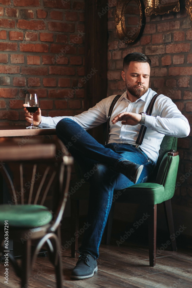 Handsome young man enjoying a glass of red wine looking at his watch Handsome young man enjoying a glass of red wine looking at his watch