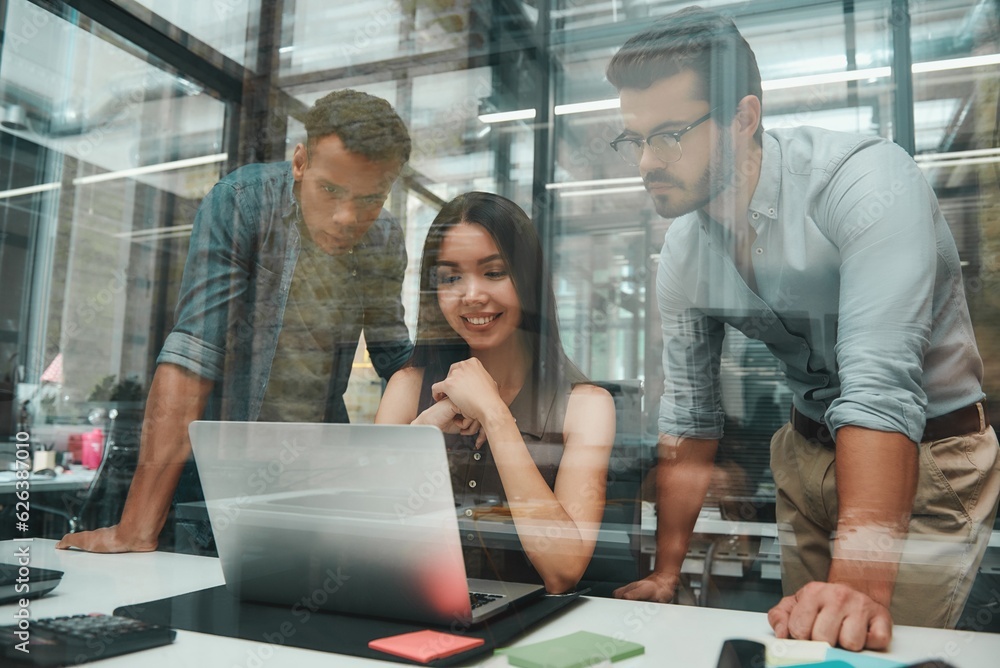 Busy day. Group of three young employees looking at screen of laptop and discussing new ideas while working in modern open space Busy day. Group of three young employees looking at screen of laptop and discussing new ideas while working in modern open space