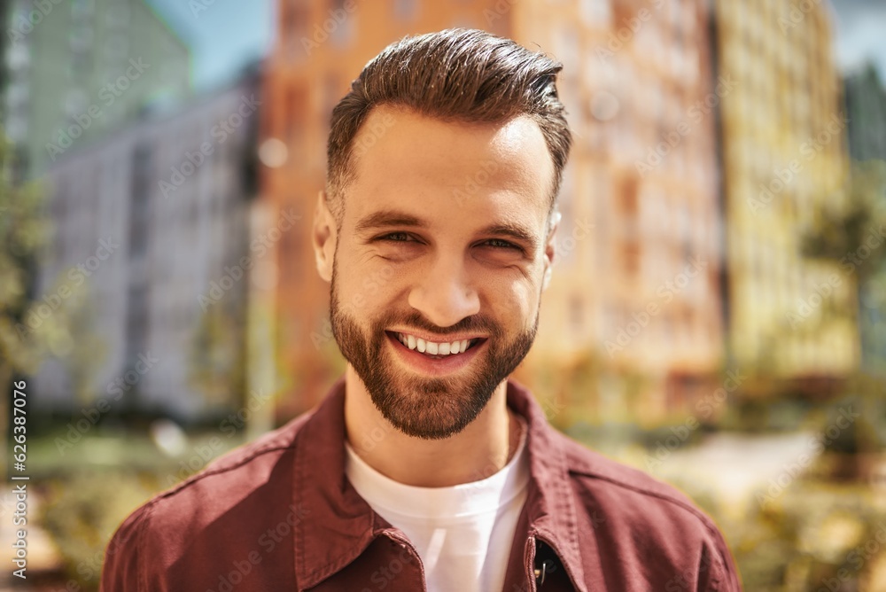 Hello Portrait of happy attractive man with stubble smiling and looking at camera while standing on the street Hello Portrait of happy attractive man with stubble smiling and looking at camera while standing on the street
