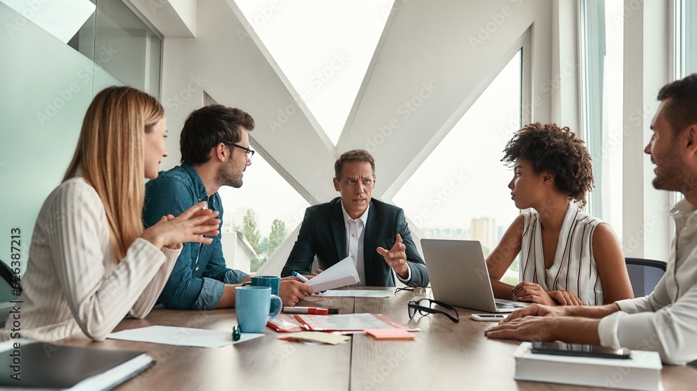 Staff meeting. Serious mature man discussing new project with his multicultural team while sitting at the office table Staff meeting. Serious mature man discussing new project with his multicultural team while sitting at the office table