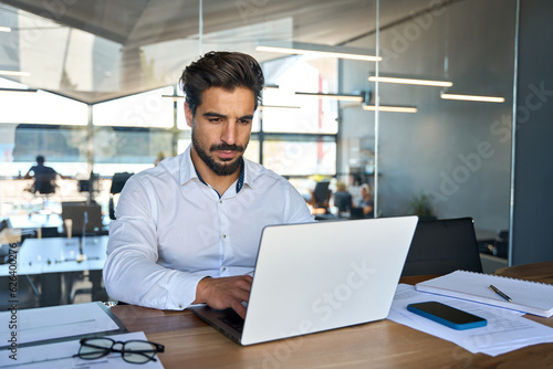 Busy young Latin business man looking at laptop at work. Focused young businessman company executive manager investor using computer sitting in office thinking of investment plan checking digital data